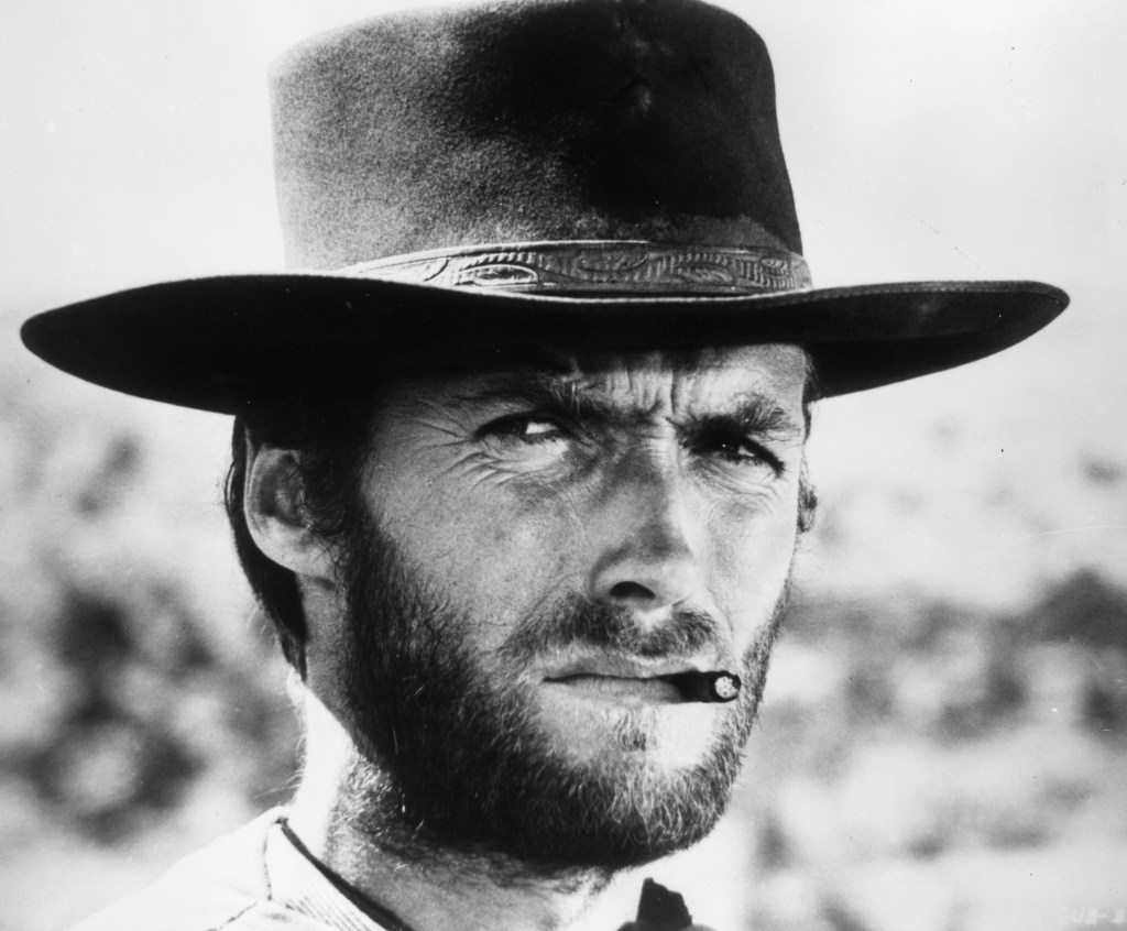 American actor Clint Eastwood squints while smoking a cigarette between his teeth in a still from director Sergio Leone's film 'The Good, The Bad, and The Ugly.' Eastwood wears a wide-brimmed leather hat. 
