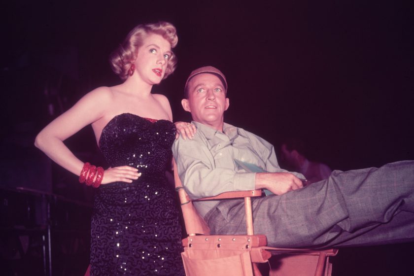 1954: American singer and actors Rosemary Clooney (1928 - 2002) and Bing Crosby (1903 - 1997) talking on the set of director Michael Curtiz's film, 'White Christmas'. Clooney is in costume in a strapless full-length black sequined gown, and Crosby wears a baseball cap and sits in a director's chair.