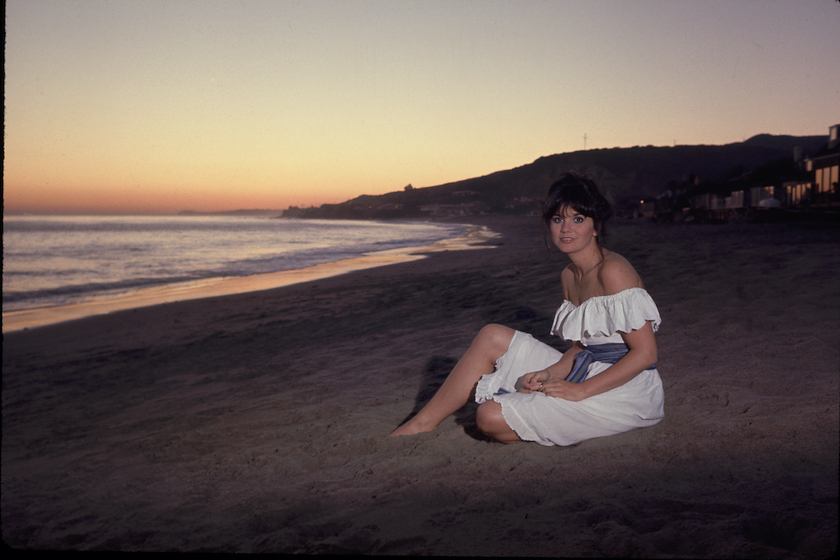 Singer Linda Ronstadt sitting on beach. 