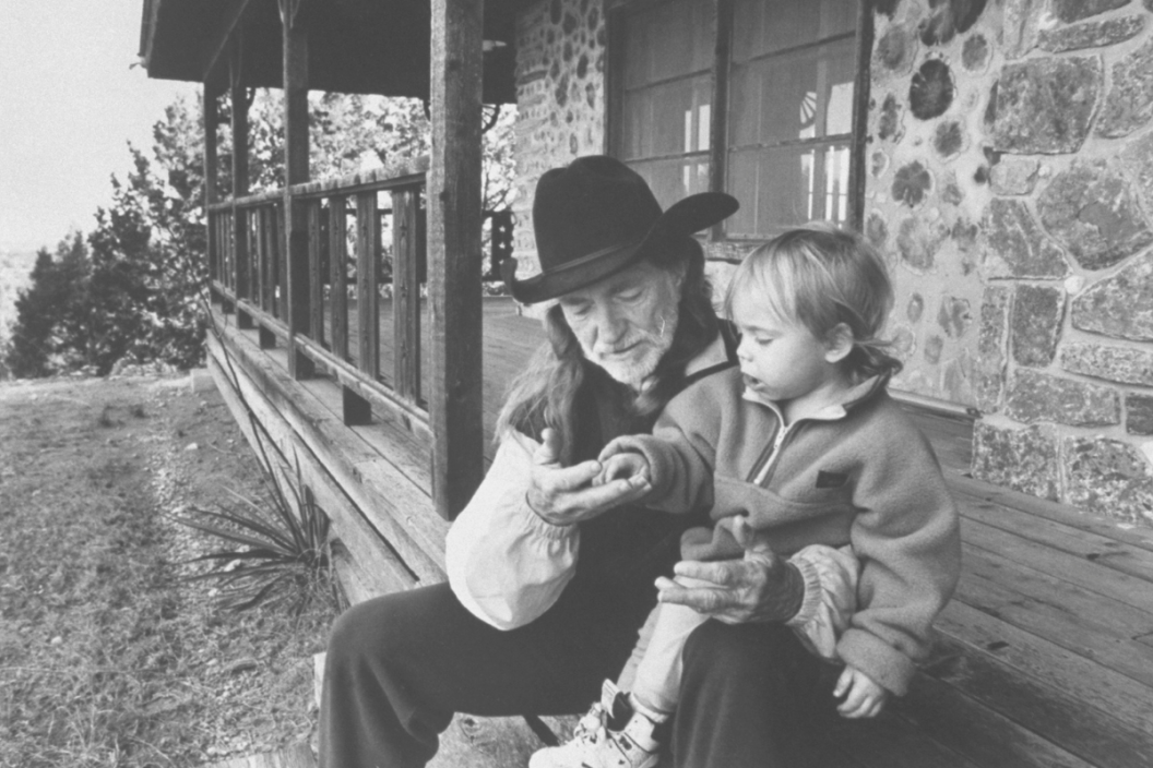 C/W singer Willie Nelson w. his son Lukas Autry, 2, chatting as they sit on front steps of his barren cabin which the IRS stripped of its furniture after it seized the 688-acre property because of his failure to pay back income taxes amounting to 16.7mi. llion