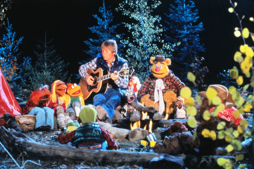 Musician John Denver during an appearance on the Muppet Show. He is seated around a campfire with (clockwise): Kermit the Frog (facing away from camera), unidentified, Janis, Scooter, John Denver, Gonzo and Chickens, Fozzie Bear and Rolf the Dog.