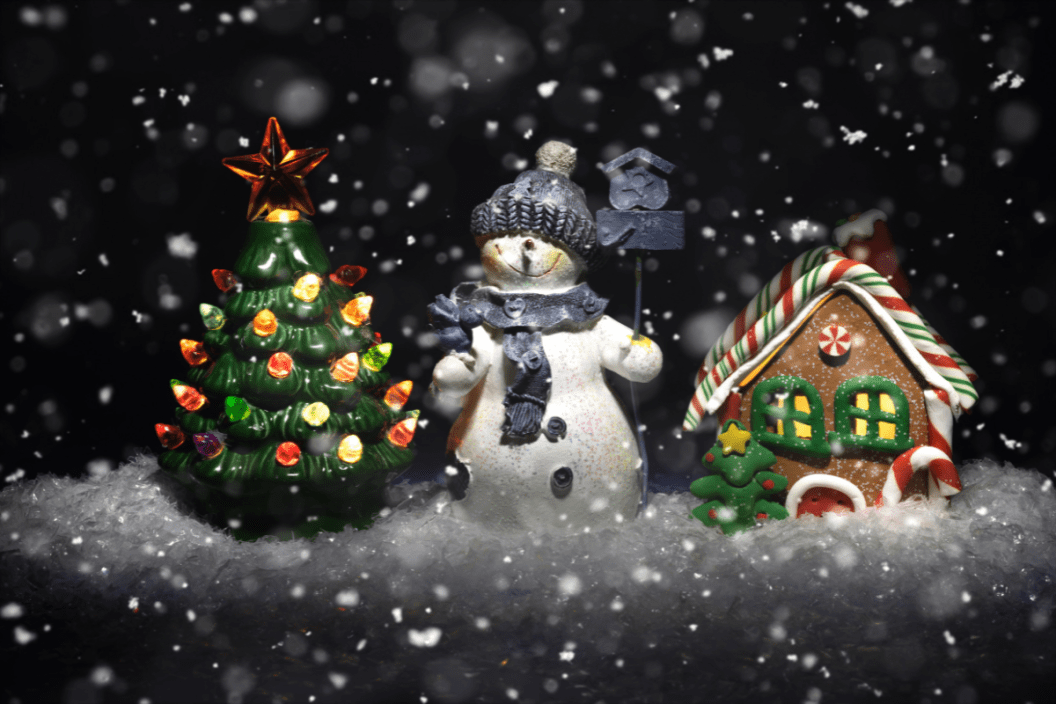A ceramic toy snowman stands in the snow between a ceramic glowing tree and a gingerbread house under a snowfall