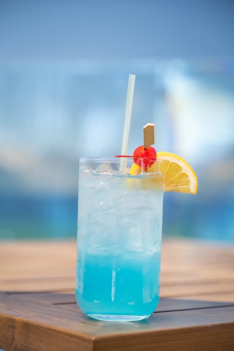 A refreshing blue cocktail on the rocks (Could be an AMF or a Blue Motorcycle or a Blue Hawaii) garnished with a lemon and a cherry sits lake side on a wooden table. The vibrant blues and greens of the lake in the background.