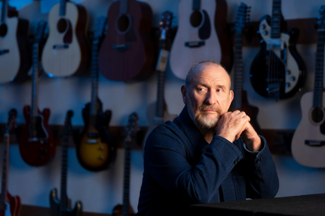 Colin Hay poses in front of wall of guitars