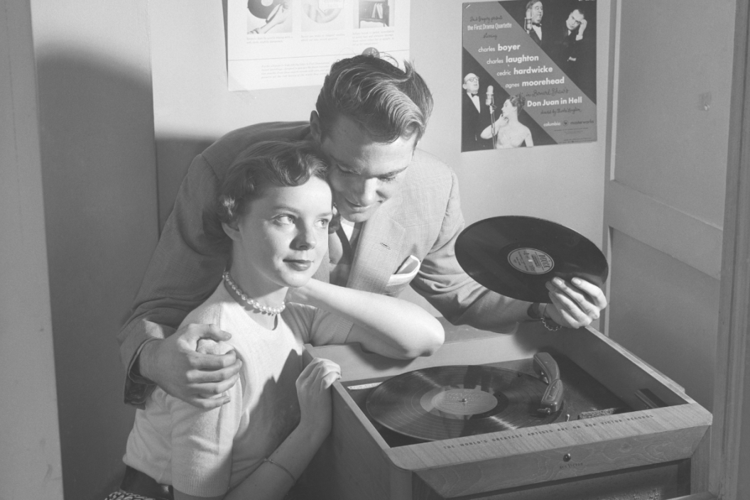 1950s COUPLE SITTING IN STORE RECORD LISTENING BOOTH MAN WITH ARM AROUND WOMAN HOLDING 78 RPM