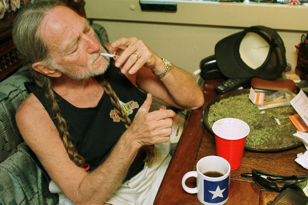 American country singer Willie Nelson takes a drag off a joint while relaxing at his home in Texas, 2000s. A large amount of marijuana is spread out on the table before him (Photo by Liaison/Getty Images)