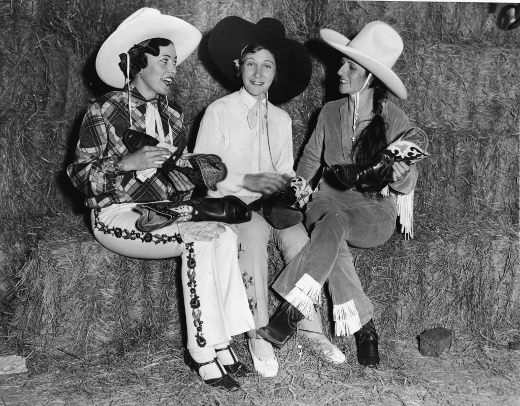 Three American cowgirls, from left, Lucyle Roberts (1909 - 1995), Alice Greenough (1902 - 1995), and Reine Shelton (1902 - 1979), sit on haybales, polishing and comparing boots in preparation for a charity rodeo in Madison Square Garden, New York, New York, October 9, 1933. 