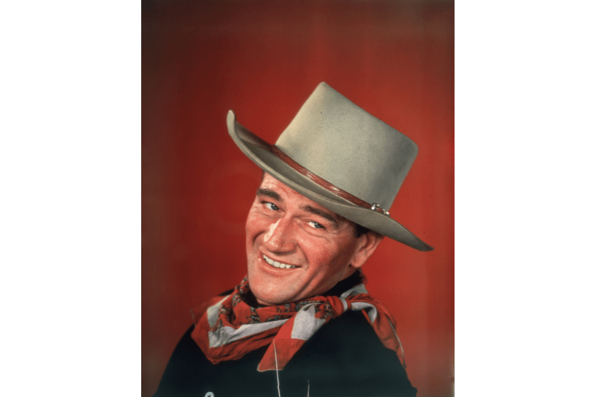 circa 1955: Studio headshot portrait of American actor John Wayne smiling in front of a red background, dressed in Western garb, with his head turned to the side
