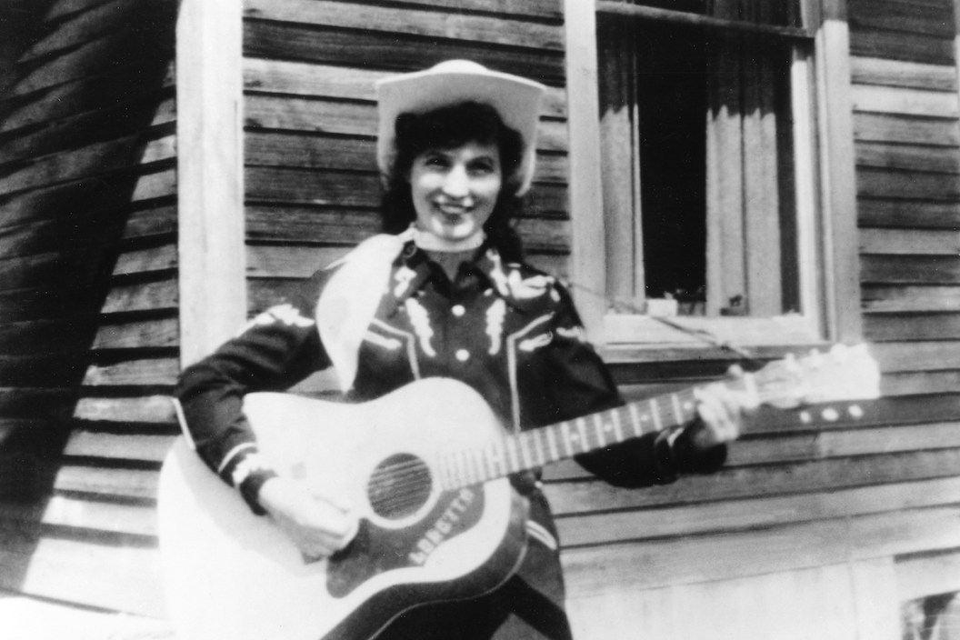 CIRCA 1960: Loretta Lynn holds her acoustic guitar as she poses for a portrait wearing a cowboy hat, a scarf and western shirt outside a log cabin in circa 1960.