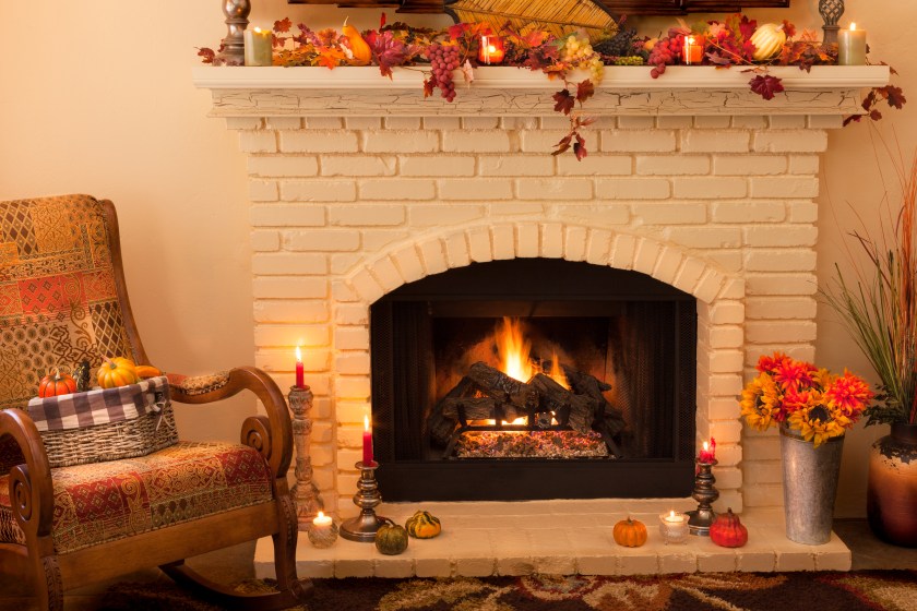 A light yellow brick old fashioned fireplace with arched opening has a roaring fire with logs and is decorated on the mantel and hearth with Thanksgiving or Autumn decorations. Candles are lit on candlesticks and fall leaves with gourds and grapes help decorate. Sunflowers and mums also fill tins and baskets on the carpeted floor. A rocking chair sits in the corner next to the fireplace in the left corner of this horizontal image. There is copy space in the left top corner.