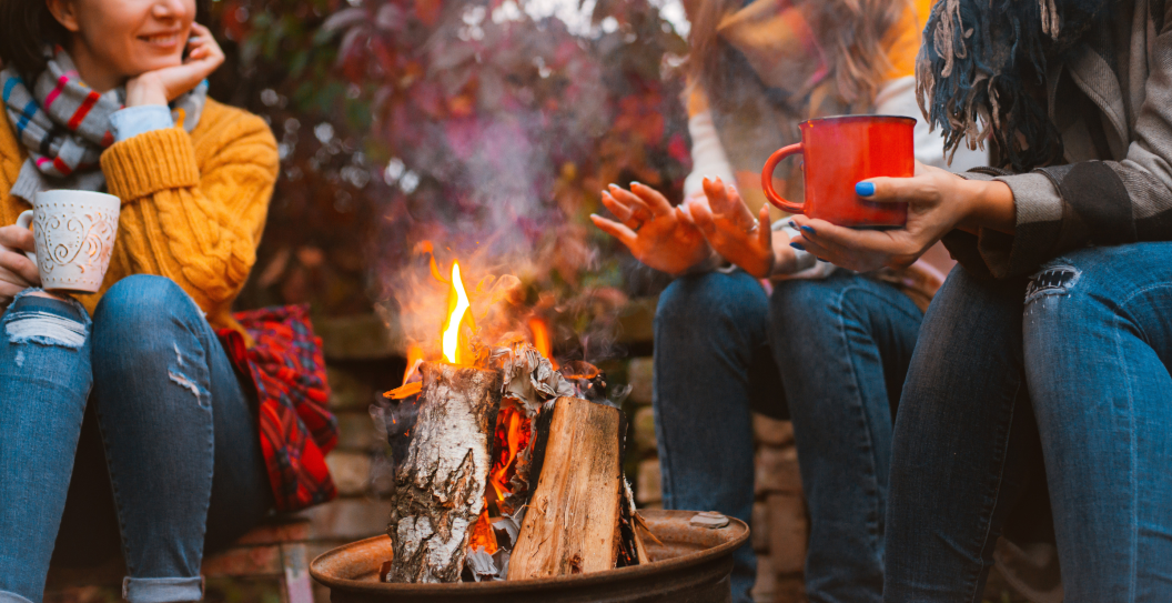 Cropped photo of three females best friends sitting around bonfire in casual clothes warming up and communicating, holding mugs with hot drinks, happy to be together outside in cold autumn evening