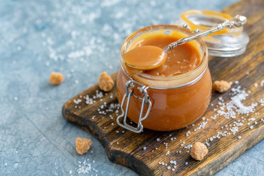 Caramel sauce with sea salt in a small jar, brown cane sugar and a spoon with salt on an old wooden serving board, selective focus.