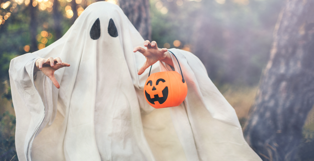 Girl wearing ghost costume holding pumpkin bucket with candies, standing in a forest on Halloween.