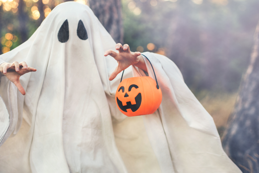 Girl wearing ghost costume holding pumpkin bucket with candies, standing in a forest on Halloween.