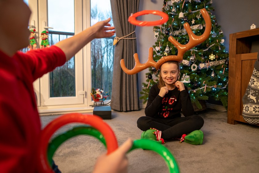 A young girl sitting at home in the living room wearing inflatable reindeer antlers. She is sitting on the floor in the living room at home opposite her brother, who is throwing inflatable rings at the antlers. The room is filled with Christmas decorations, ready for Christmas Day.