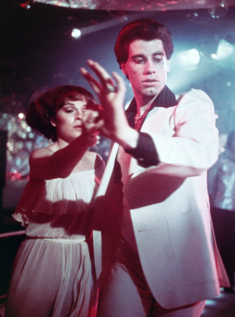 American actors John Travolta, wearing a white suit with a black shirt, and Karen Lynn Gorney disco dance in a still from the film, 'Saturday Night Fever,' directed by John Badham, 1977. 