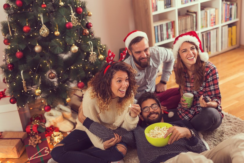 Group of young friends sitting on the floor next to a Christmas tree, eating popcorn and watching a Christmas movie. Focus on the girl wearing antlers
