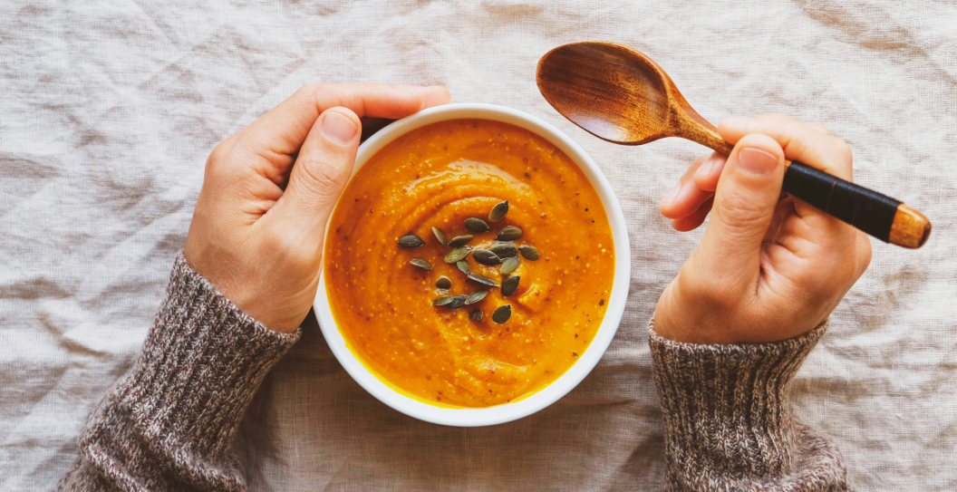 Male hands holding a bowl of hot pumpkin soup served with squash seeds on white tablecloth background, from above. Flat lay. Homemade autumn food. Popular Thanksgiving dish.