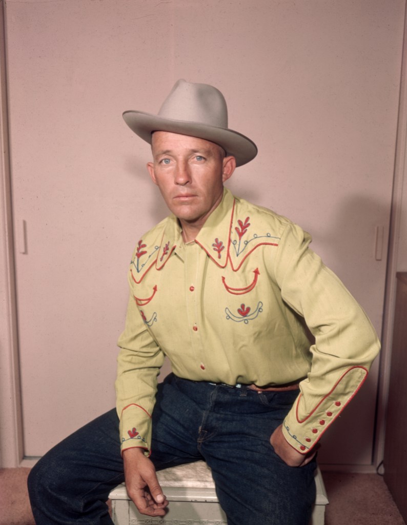 Studio portrait of American actor and singer Bing Crosby seated with his legs spread apart and his fist on his hip, wearing a western shirt, jeans, and a cowboy hat.
