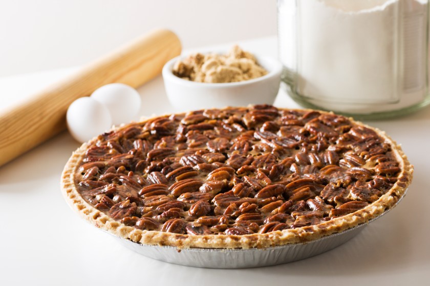 A freshly baked pecan pie with ingredients and a rolling pin in the background. The delicious homemade dessert is ready for a holiday or other special occasion.