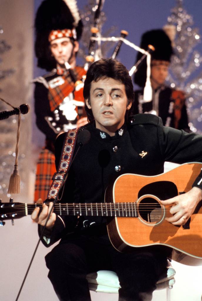 Paul McCARTNEY, in Wings, performing on TV show, playing acoustic guitar, with bagpipe players.