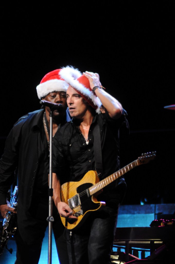 Bruce Springsteen and Clarence Clemons performing on stage wearing Christmas hats.
