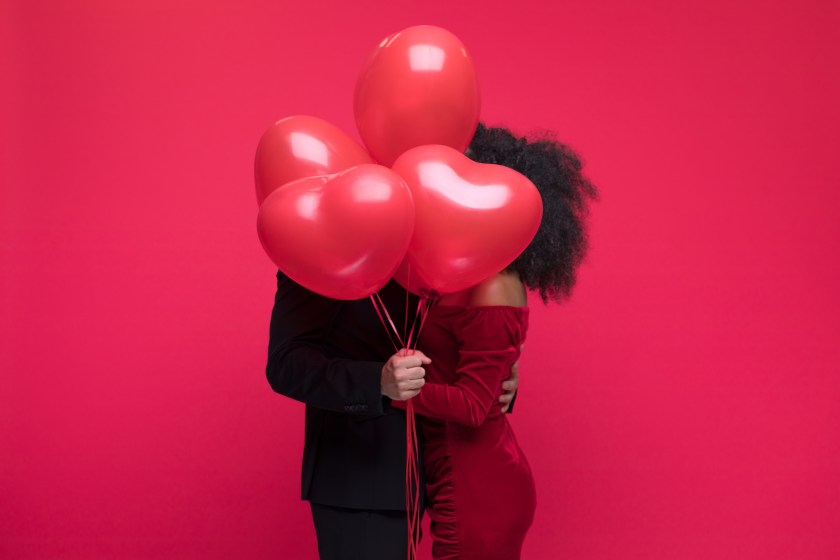 Young couple wearing elegant covering with bunch of heart shaped balloons. Studio shot, red background.