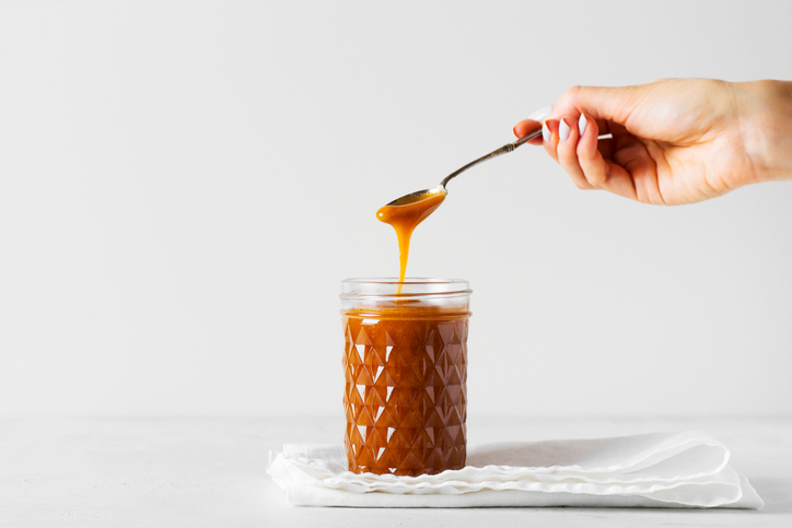 Liquid brown caramel in glass or jar with woman hand holding a spoon on white background, copy space