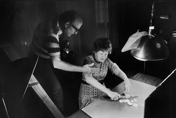 Chef Julia Child chopping squash as her husband, Paul photographs her for an upcoming cookbook. 