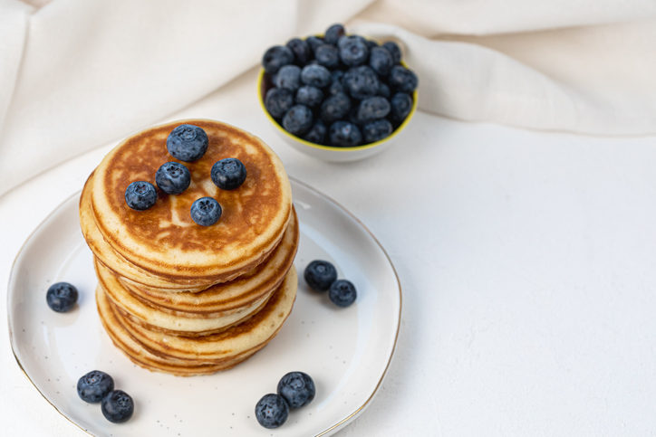 Stack of delicious pancakes on white plate with fresh blueberries on and in bowl